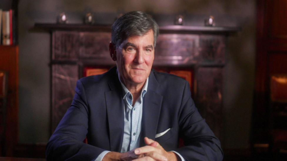 A man with short dark hair, which is turning grey, is sat in front of a large dark fireplace. He is wearing a navy suit jacket and blue, open collar shirt. His hands are clasped in front of him as he rests his elbows on a table.