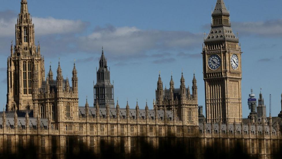 A view of Big Ben from a greater distance with the gothic-looking Parliament also seen to the left of the Elizabeth Tower. Big Ben towers over the surrounding buildings. The sky is mostly blue with a few grey clouds. There is a dark shadow covering the bottom of Parliament.