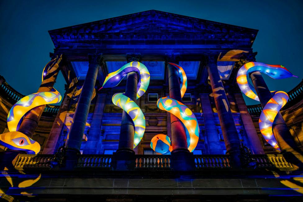 Several large, brightly coloured serpent sculptures illuminated from within coil around the columns of a grand historic building during the Paisley Halloween Festival. The building façade is bathed in blue light, while the glowing snakes feature vivid yellow, orange, green and blue patterns
