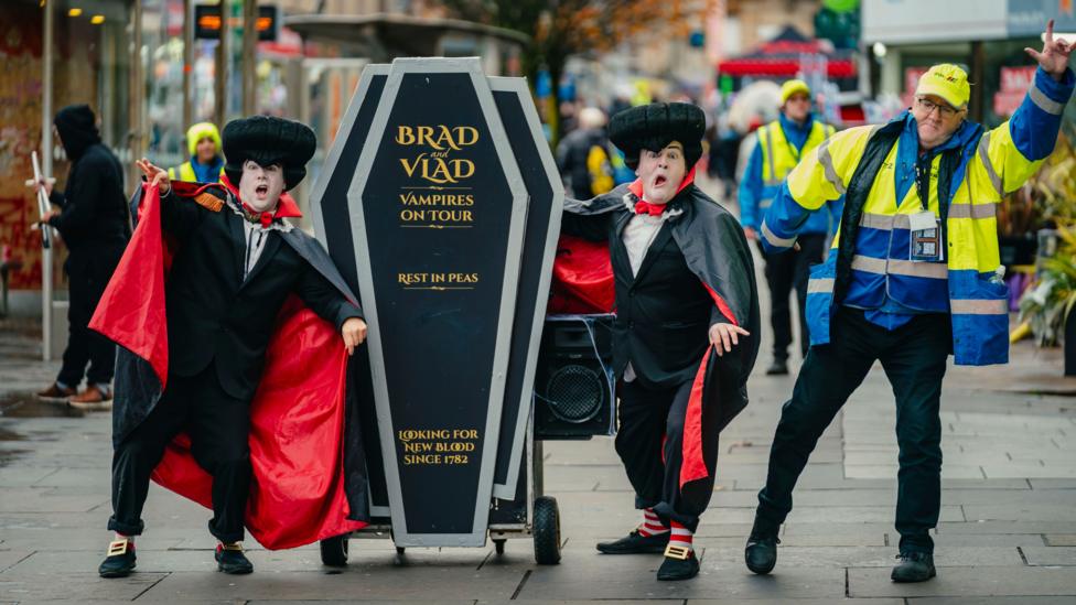 Two performers dressed as vampires pose beside a coffin prop on a city street during the Paisley Halloween Festival, while event staff and festival-goers look on in the background.