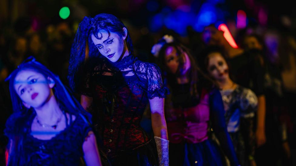 Several participants wearing gothic-style Halloween costumes and theatrical face paint take part in the Paisley Halloween Festival parade at night. A central figure in a black and red outfit is highlighted by blue stage lighting, while other costumed participants and colourful festival lights create a blurred backdrop