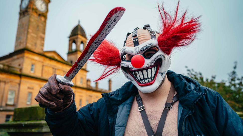 A costumed participant wearing a dramatic clown mask with exaggerated teeth, a red nose and bright red hair poses with a large prop blade during the Paisley Halloween Festival. The historic Paisley Town Hall clock tower and surrounding architecture are visible in the background.