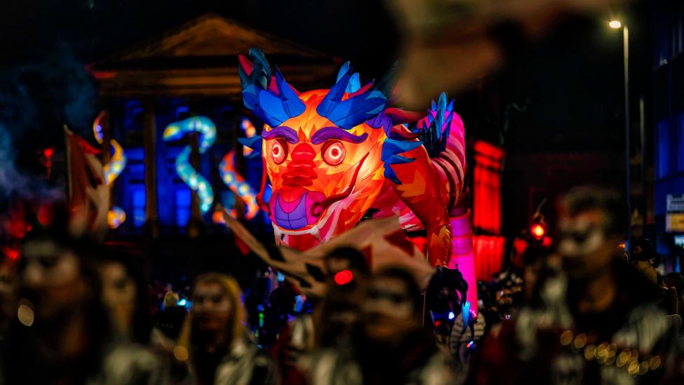 A colourful giant lantern sculpture resembling a mythical creature moves through the Paisley Halloween Festival parade at night. The lantern is lit from within and features bright orange, pink and blue geometric patterns, while costumed performers appear in the foreground and historic buildings are illuminated with vivid blue and red lighting in the background. Crowds line the street