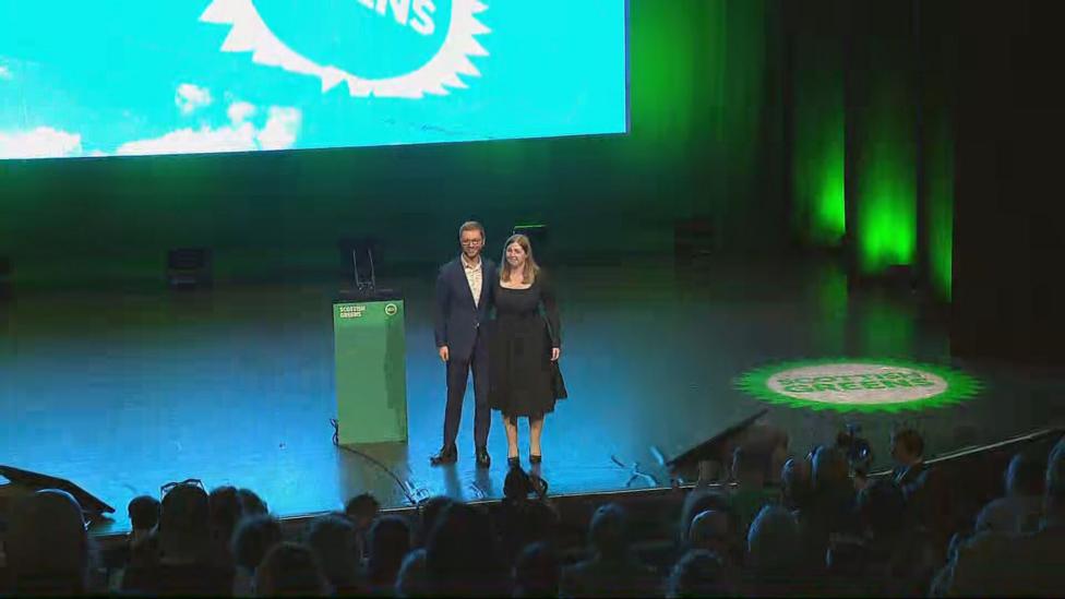 Ross Greer and Gillian Mackay stand on stage in front of a large Scottish Greens conference screen while attendees watch from the auditorium during a party conference event.