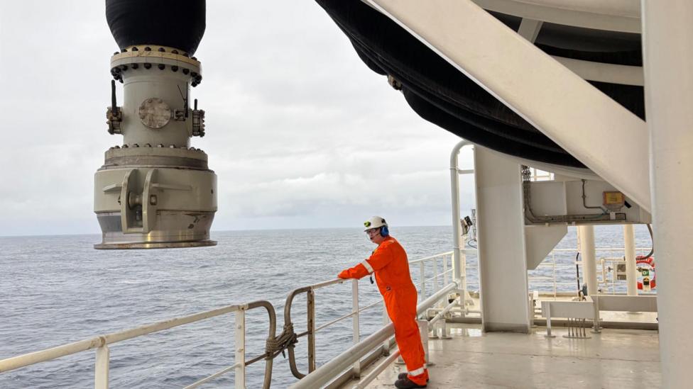 Man in orange overalls and a white helmet leans over a railing with the sea beyond. There is a large nozzle hanging down near him