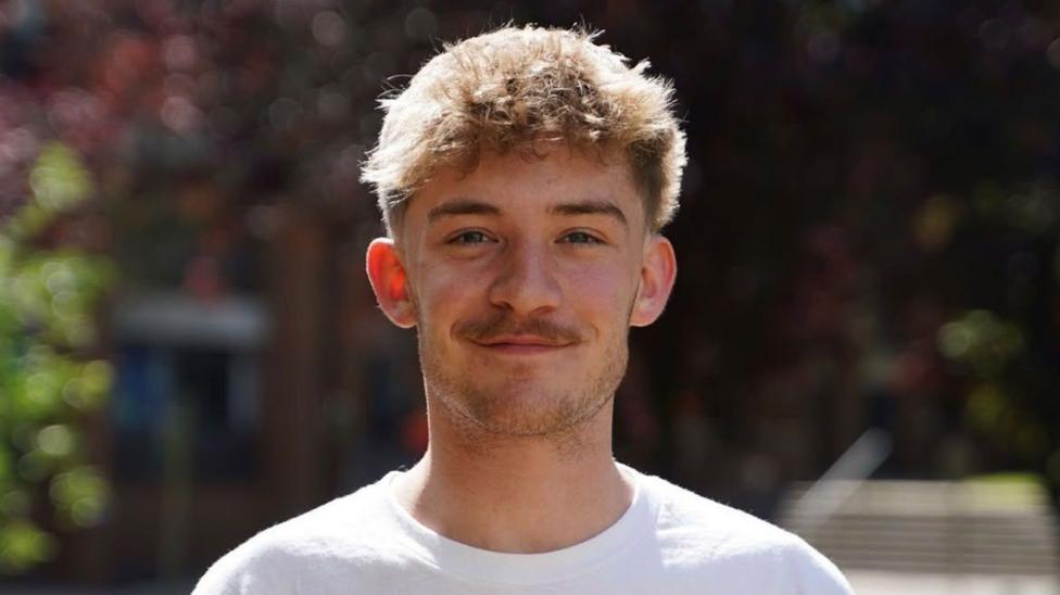 Head and shoulders shot of Lewis Wilson, a young man in a white T-shirt, with a moustache and light beard