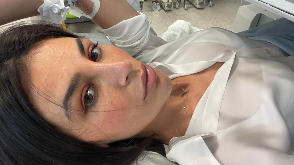 Carla lying on a hospital bed. She has long brown hair and brown eyes. She is wearing a white shirt and gold necklace. 