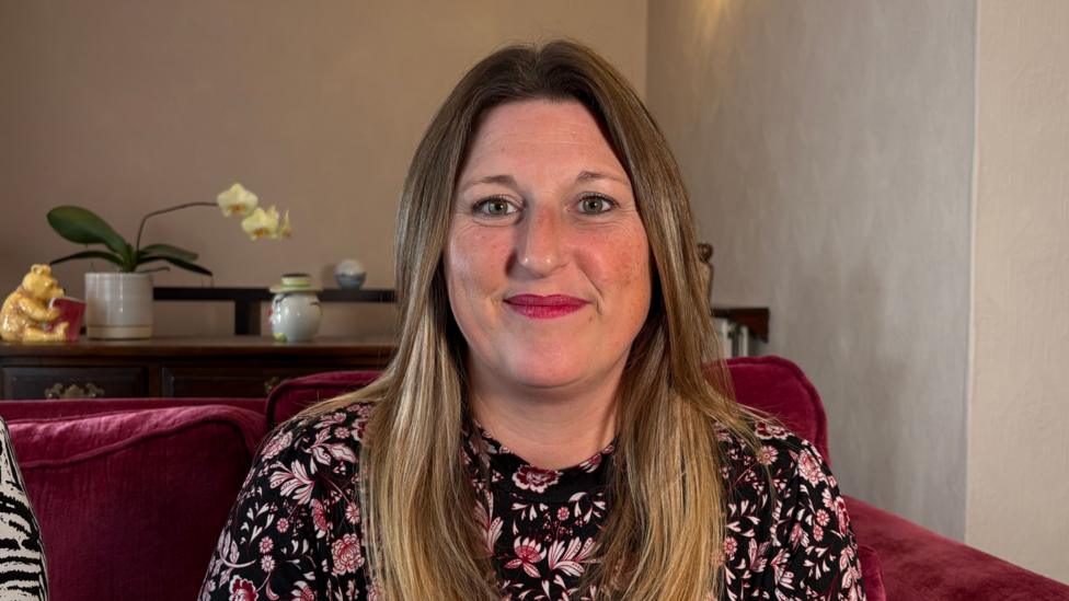 Jessica Taylor looking directly at the camera. She is sitting on a dark red sofa in a room with cream-coloured walls. She is wearing a flowery-patterned top and is smiling, though isn't showing her teeth. She has straight hair that is falling over her shoulders. Behind her is an orchid in a pot on a table, with other knick-knacks next to it. 