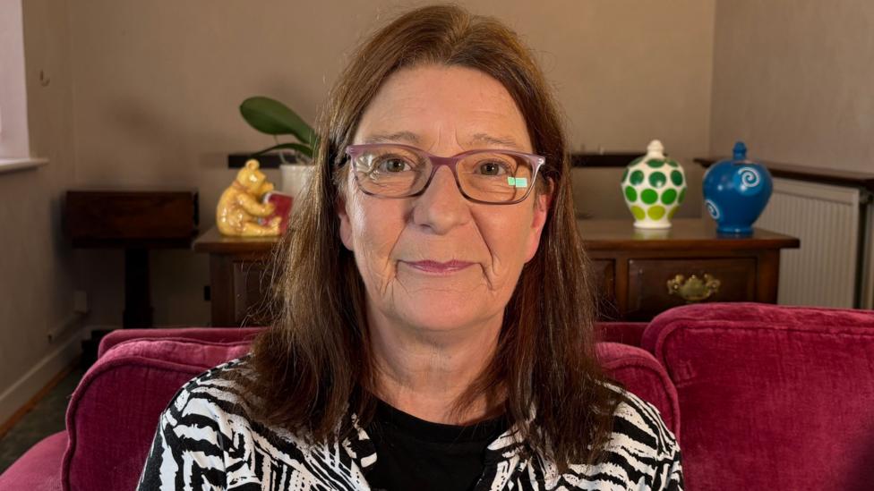 Karen Badenoch looking directly at the camera. She is sitting on a dark red sofa in a room with cream coloured walls. She is wearing a black and white patterned top with a round-necked black T-shirt underneath. She has rectangular-shaped glasses on and straight brown hair that is falling to just below her shoulders. Behind her is a desk with several money boxes on, including one in the shape of Winnie the Pooh. 