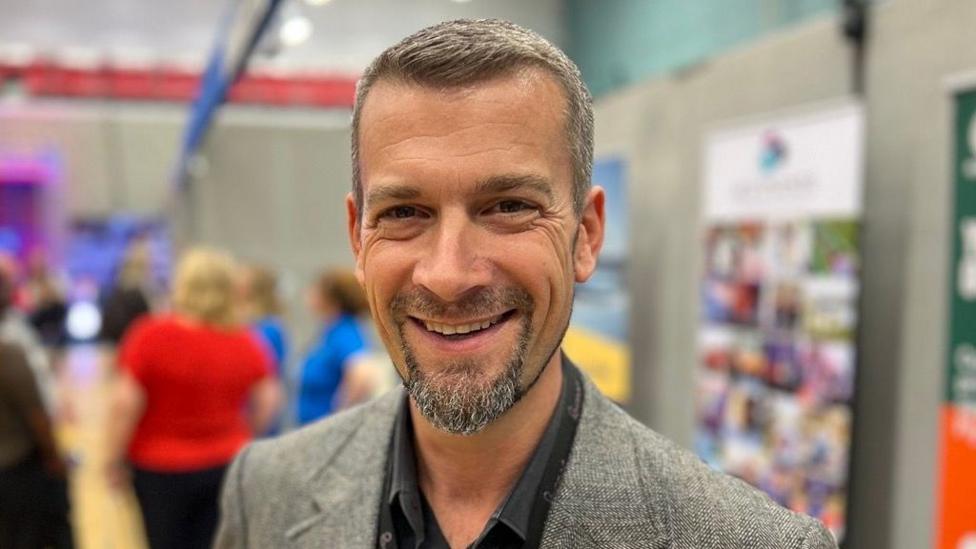 Pete Adams is wearing a black shirt and a grey fine check blazer and teh background is blurred
