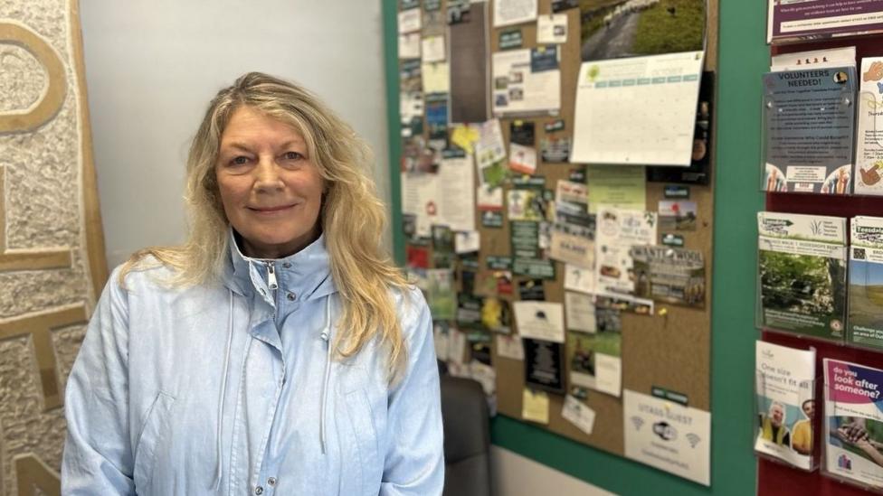 A woman is standing inside in front of a notice board, she has long blonde wavy hair and is wearing a pale blue zip up anorak. 