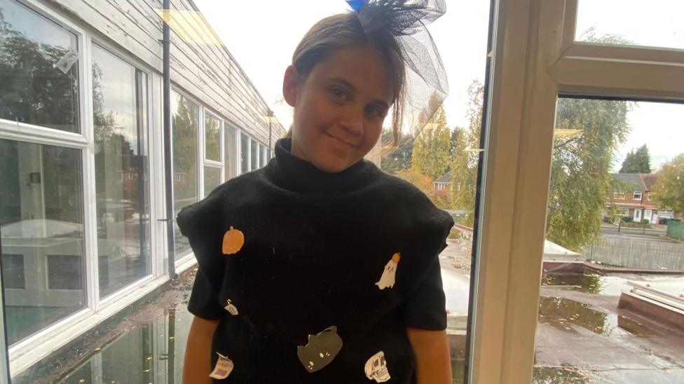 Child wearing a black homemade Halloween costume decorated with hand-made pumpkins, ghosts and spooky shapes, standing in front of a large window.