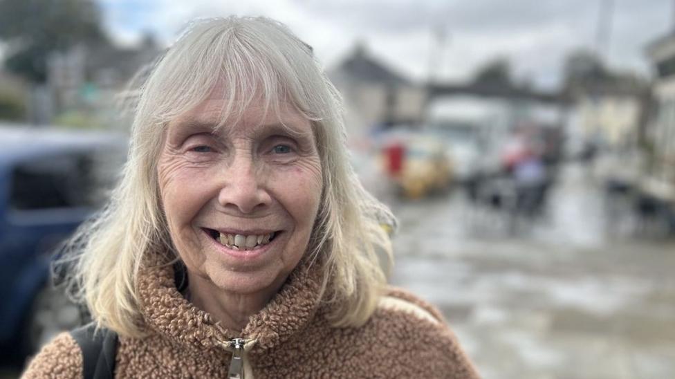 A woman is standing on the high street in Middleton-in-Teesdale, the photo is in portrait mode so the background is blurred. She has shoulder length grey-blonde hair with a fringe. She is wearing a borg style tan coloured fleece. 