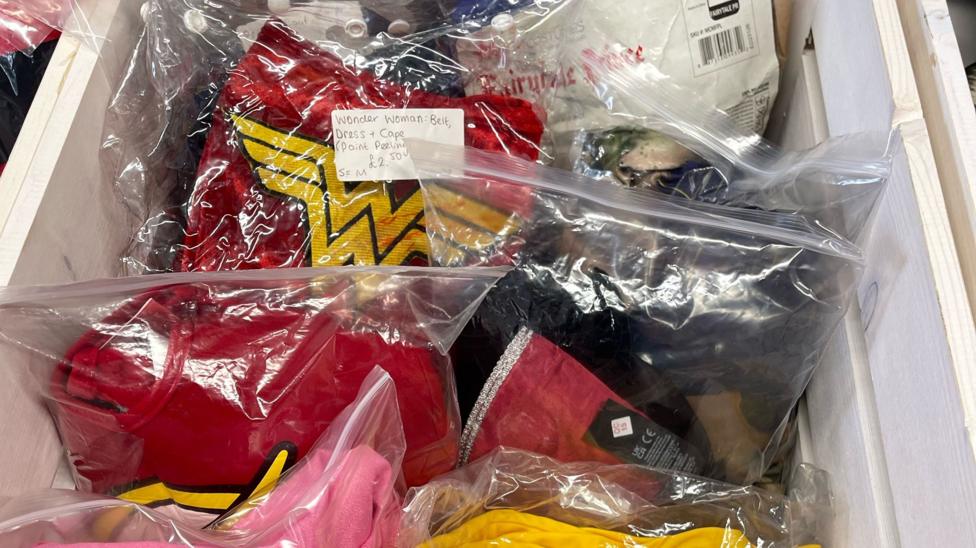 A wooden display drawer filled with second-hand children's Halloween costumes stored in clear plastic bags, each labelled with a low sale price.