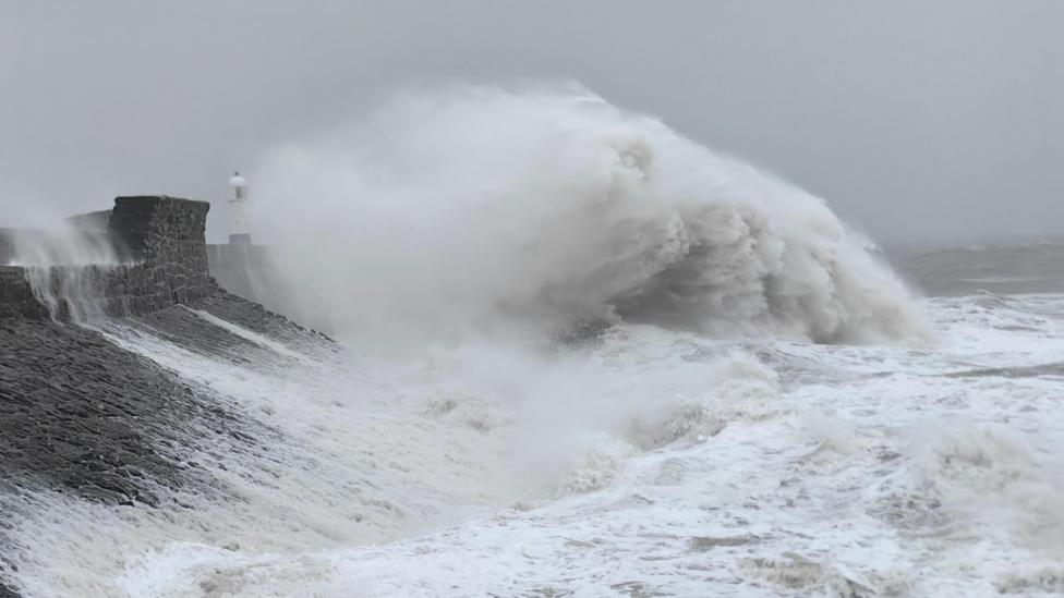 A giant wave is crashing on to a sloping sea wall. A lighthouse can be seen in the distance.