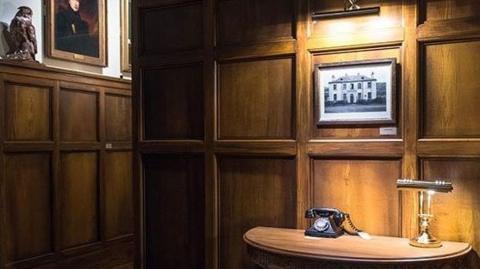A room with wood panelled walls with a framed black and white photograph of Ballechin House on one wall. Below it is a small table with a telephone and a lamp.
