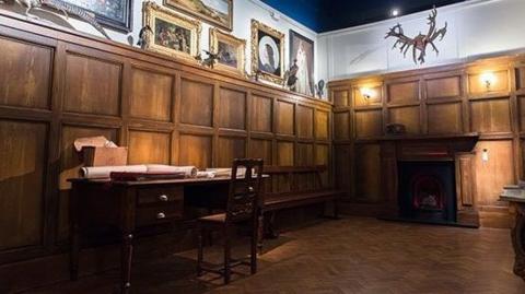 A room with wood panelled walls, a wooden desk and chair and ornately framed pictures on the walls above the panelling.