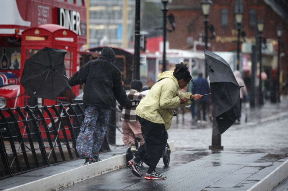A woman, wearing a yellow coat and black trousers, struggles to hold onto her umbrella during strong winds in Liverpool