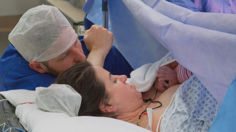 Kate is lying on a hospital bed with her newborn daughter on her chest. Matt is crouched down next to her and they are both staring at their daughter.