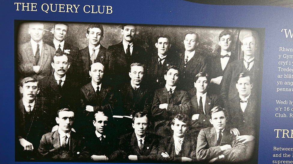 A black and white photograph of the Query Club members taken in the 1920s has been incorporated into an information panel. There are 20 men all dressed in suits and ties pictured in three rows. Above the photograph the words "The Query Club" are printed, and to the right some bits of words, mostly in Welsh, can be seen.