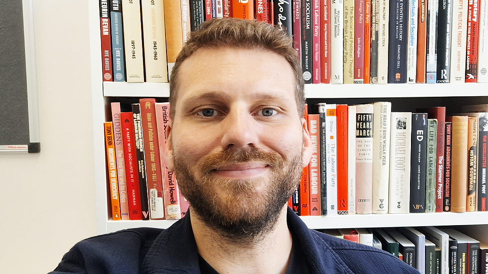 Head and shoulders shot of Nye Davie, who is a white man in his 20s or 30s. He has short light reddish-brown hair and a beard. He is looking directly at the camera and smiling. Behind him are white bookshelves 