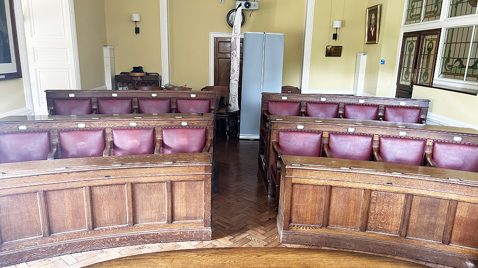 The town council chamber in Bedwellty House. There are two curving rows of eight seats each, which have reddish-purple leather padded backs with a small plaque on the top of each backrest and a pew-like writing space in front. There is an aisle in between separating them into blocks with four seats on either side. To the right are stained glass obscured internal windows and a door. There is a wall-mounted projector at the back, a tall display poster turned back to front and two mounted wall lamps either side of the room. A photograph of former Labour leader Sir Neil Kinnock is on the side wall.