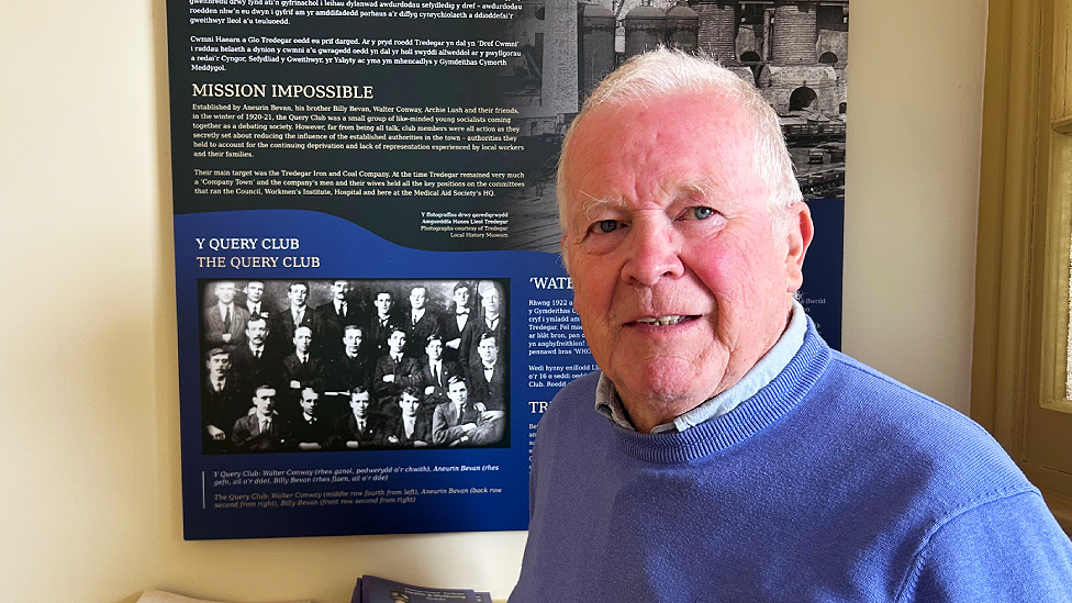 John Morgan is a white man in his 70s. He has short white hair and a ruddy complexion. He wears a peacock blue jumper over a light blue shirt collar. He is standing to the side of the Query Club photograph and information panel.