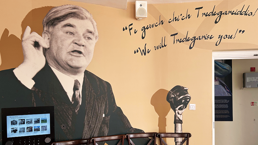An image of Aneurin Bevan speaking into a microphone has been enlarged and displayed on one wall of the Tredegar Medical Aid Society Heritage Centre. He has a finger raised mid-speech. The works "We will Tredegarise you!" and the Welsh equivalent "Fe gewch chi'ch Tredegareiddio!" and written in cursive script nearby. An interactive screen is visible in front of the image, and an open door leading to a hallway on the right of the picture.