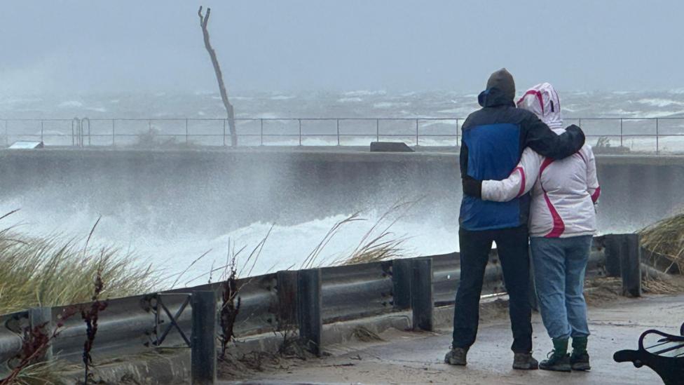 A couple huddled together looking at waves crashing at the entrance to Nairn harbour.