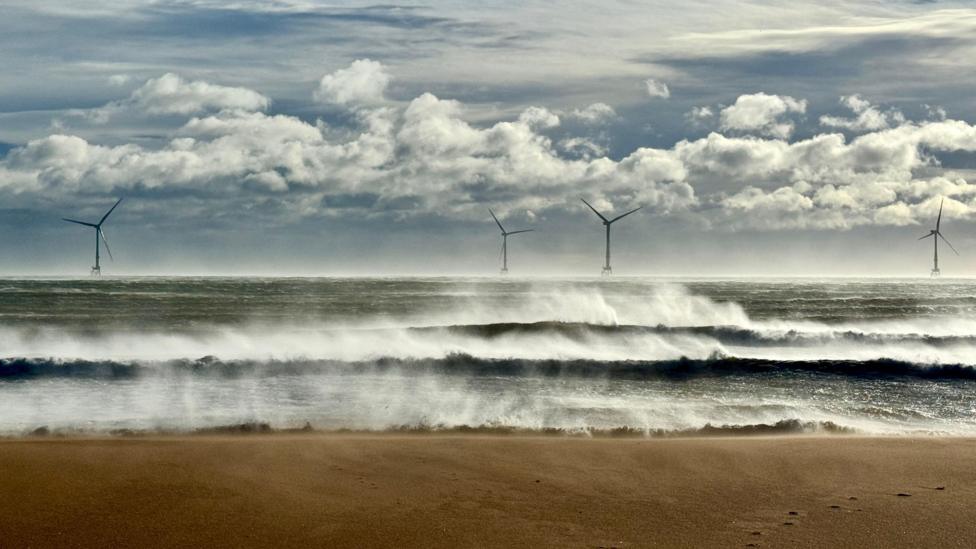 Spray is blown back of the tips of waves coming in at Balmedie beach. It is a bright day. There are offshore wind turbines on the horizon.