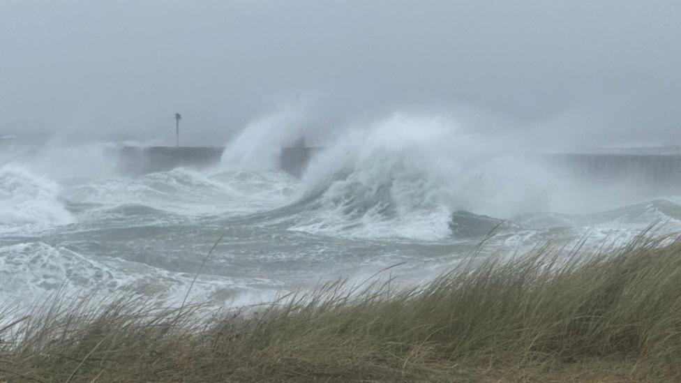 The rough sea is viewed across the grassy top of a sand dune.