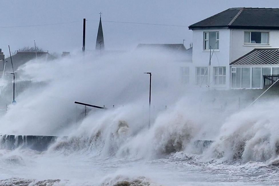 The waves are white a frothy as the water flies up and over a wall near a row of houses.