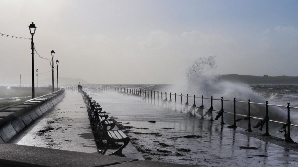Seaweed and other material lies strewn across a wet path next to benches as waves smash into a sea wall at Largs.