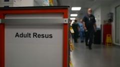 A trolley is seen in a corridor. It's in the foreground of the picture with the words: Adult Resus. Three members of staff can be seen out of focus on the righthand side