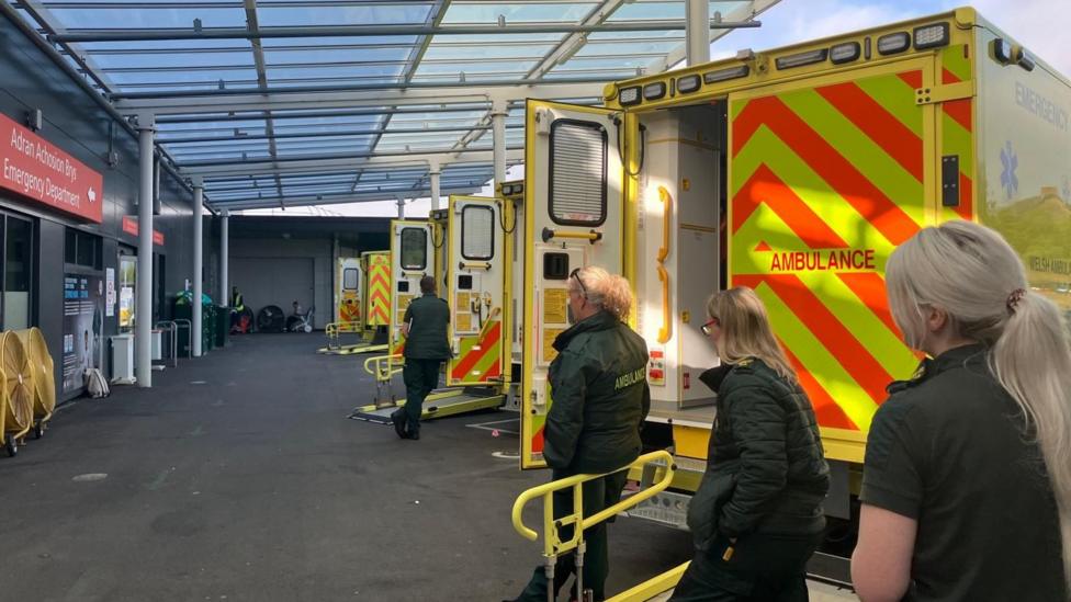 Four paramedics and ambulance technicians stand with their backs to the camera with their arms crossed or in their pockets next to their vehicles. They are parked outside the hospital's emergency department.