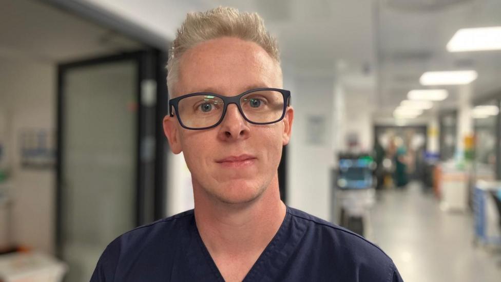 A young man smiles to camera. He has short cropped blonde hair and black rimmed oversize glasses. He wears dark navy v-neck scrubs.