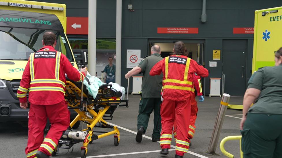 Two paramedics and three members of the air ambulance wearing red and yellow uniforms push a patient in a trolley towards the doors of accident and emergency. Two ambulances are parked either side of the action.