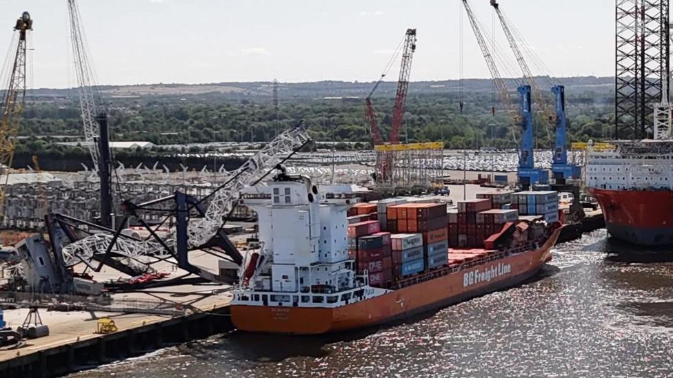 Aerial view of a container ship at a dock. It is a low-lying, orange coloured vessel, its deck stacked with containers. A crane lies crumpled next to it with the top part resting on the vessel's control tower.
