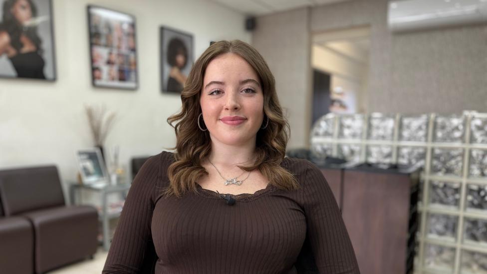 Trearna Hart is a young white woman with long brown wavy hair. She wears a long sleeved brown top. Behind her is a glass screen, behind the screen is where customers have their hair washed. On the wall behind her are framed prints of hair models.