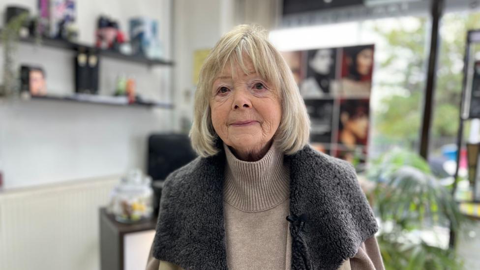 Dorothy Senior stands in M&M Spa, she is white with a grey bob and fringe. She wears a brown jumper and a brown coat. Behind her is the till and the shop front window.