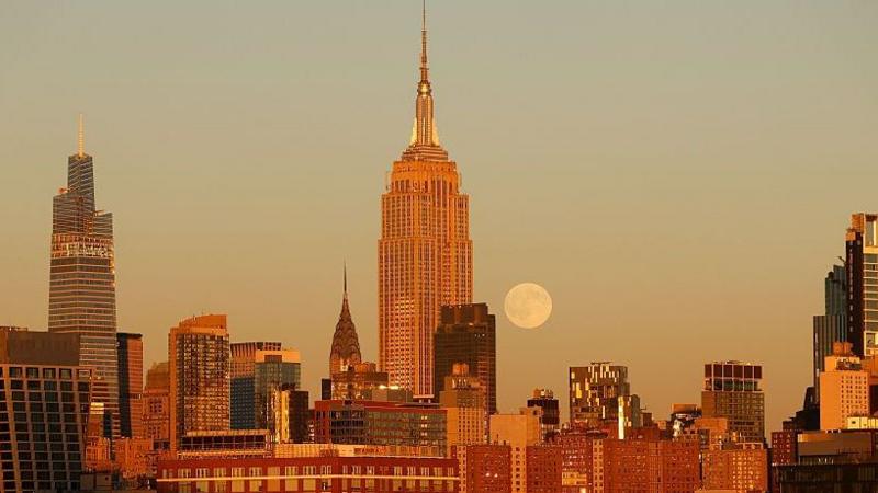 The full moon rises in an a dusty-yellow and blue sky over the city skyline, which includes New York City's Chrysler Building and the Empire State Building.