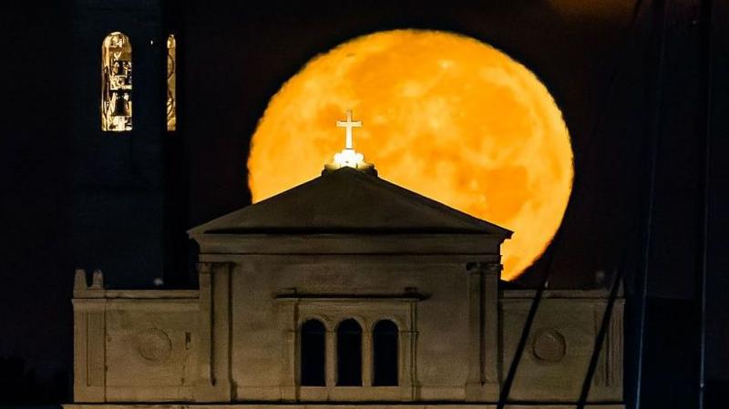 A huge yellow moon rising behind a church with a gold cross at the top of it.
