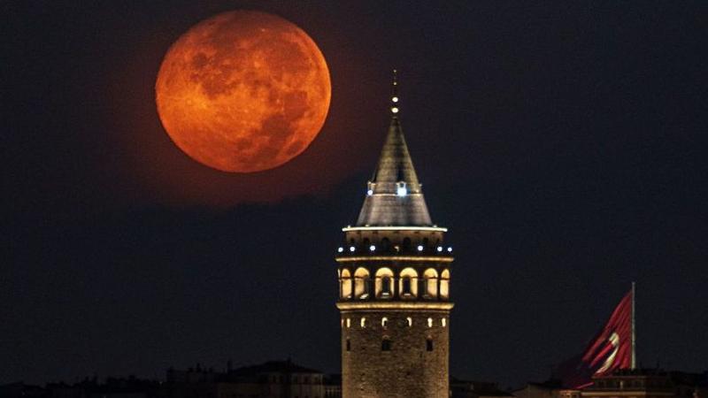 A large orange full moon in the night sky glows over a coned-top tower. There are lights on in the tower's arches and the Turkish red and white flag is next to it.