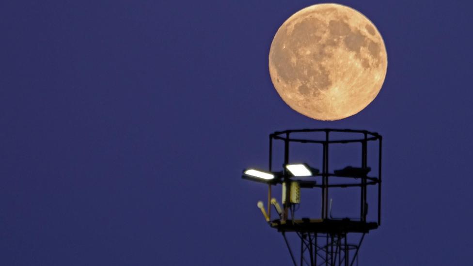 A large, glowing yellow moon sits above a metal round structure with lights on it. The moon looks like it's going to sink down into the structure - like a basketball heading into a net.