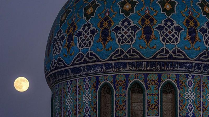 The full moon looking small in the sky compared to the large dome of 17 Ramadan Mosque which has colourful and intricate patterns on it.