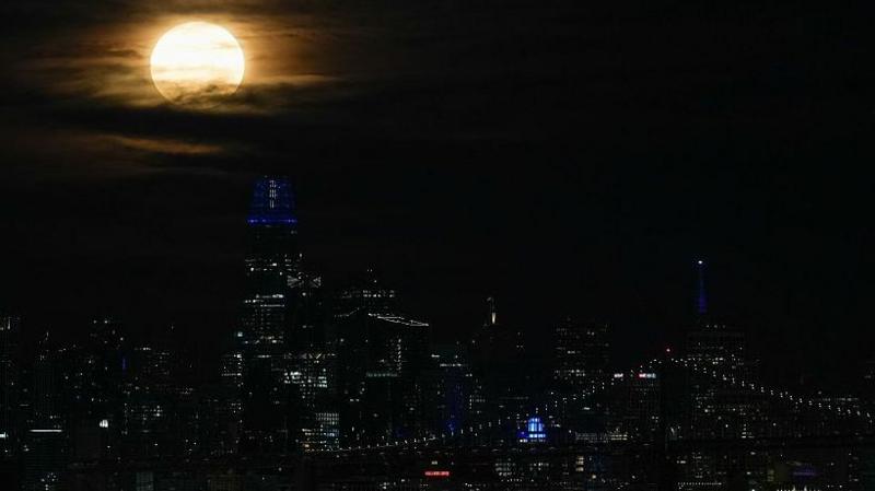 San Francisco's sky scraper lights and the lights of a bridge look dim compared to the powerful light of the full moon in the cloudy sky above them.