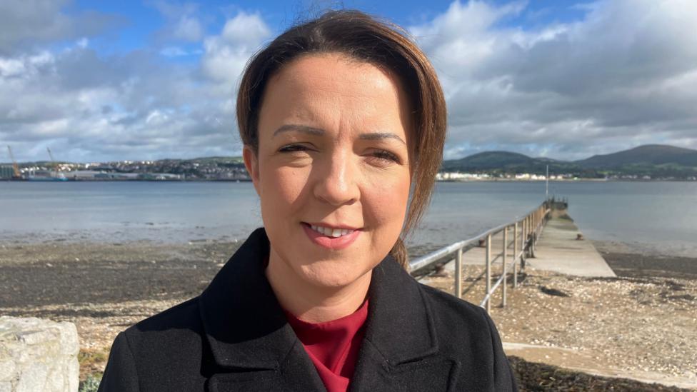 A woman with dark hair smiles at the camera, she is wearing a black coat and red top