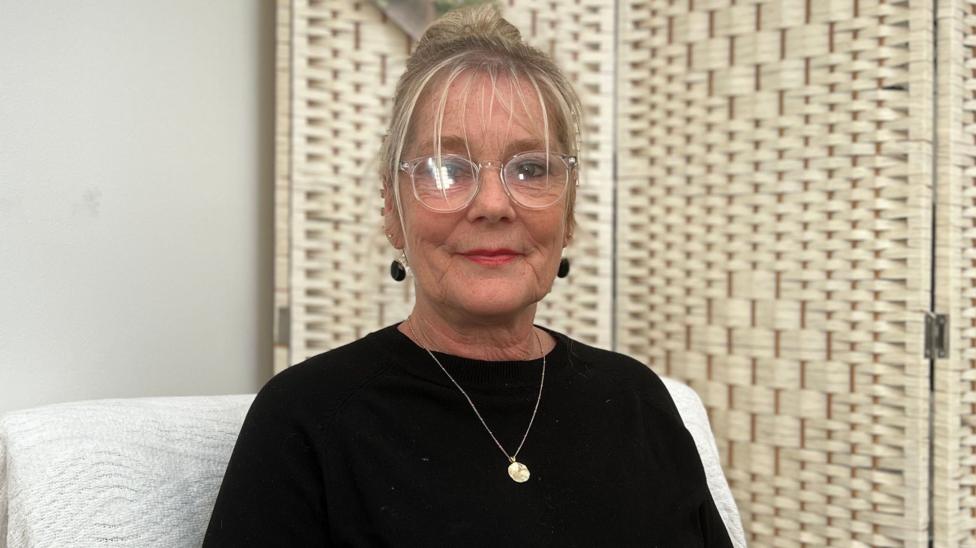 Sally Agathangelou is seated in an upholstered chair, wearing a black long-sleeved top, glasses and a necklace. A woven folding screen stands behind the chair against a plain wall, with soft indoor lighting throughout the room.