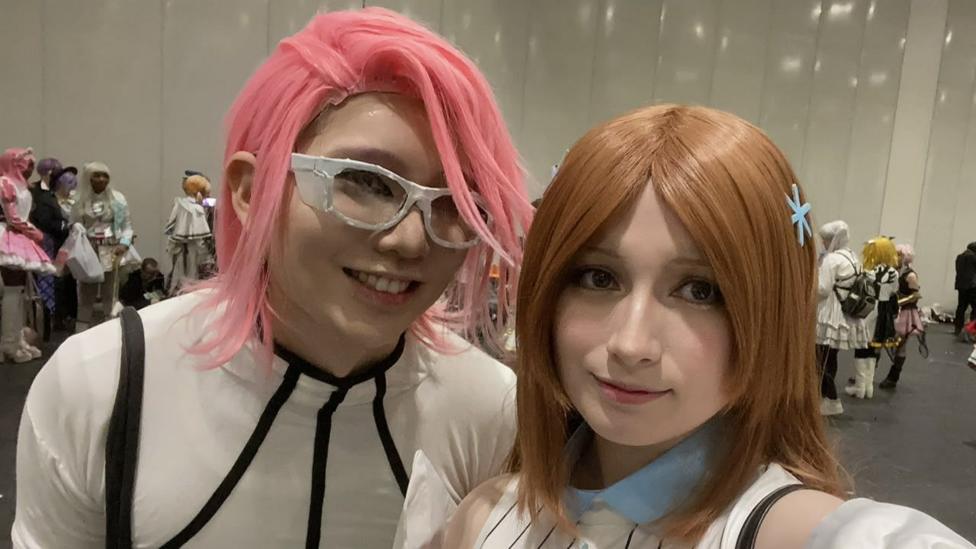 Nicholas on the right wears a white bodysuit with black piping and a pink wig. Ruby wears a blue collar and white striped outfit and a brown wig. They both look at the camera and smile.