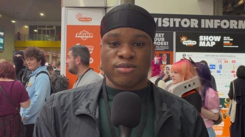 Itanna Alakija stands facing the camera and smiles as fellow Comic Con attendees mill around in the background. He wears a black protective cover on his hair, a green jumper and black jacket.